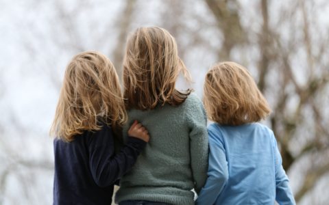Kolme pitkähiuksista lasta seisoo selin kameraan, samalla toisistaan kiinni pitäen. vasemman puoleisella lapsella on yllään musta paita, keskimmäisellä vihreä ja oikeanpuoleisella vaalean sininen.