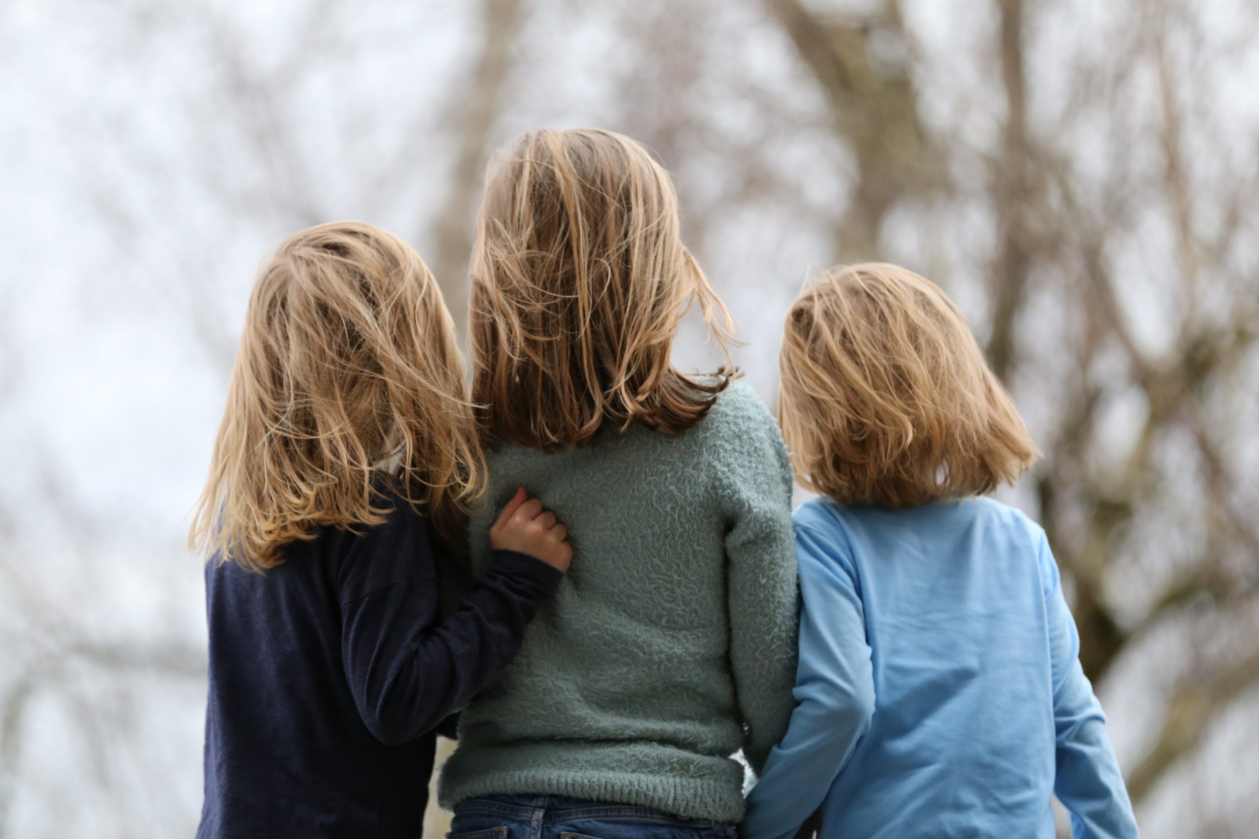 Kolme pitkähiuksista lasta seisoo selin kameraan, samalla toisistaan kiinni pitäen. vasemman puoleisella lapsella on yllään musta paita, keskimmäisellä vihreä ja oikeanpuoleisella vaalean sininen. Lasten hiukset hulmuavat tuulessa.