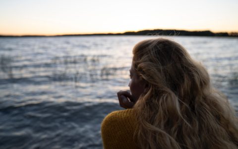 vaaleahiuksinen nainen istuu selkä kameraan päin ja katsoomerelle tai järvelle. Ilta alkaa hämärtyä ja vedessä on pientä aaltoilua. Naisella on yllään villapaita