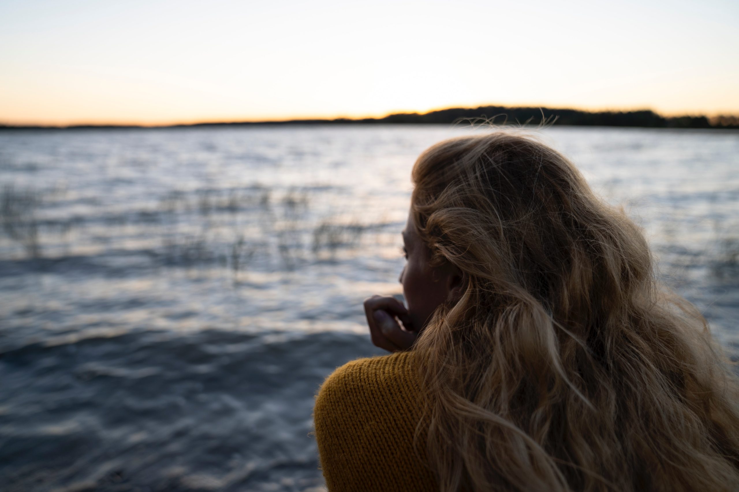 vaaleahiuksinen nainen istuu selkä kameraan päin ja katsoomerelle tai järvelle. Ilta alkaa hämärtyä ja vedessä on pientä aaltoilua. Naisella on yllään villapaita