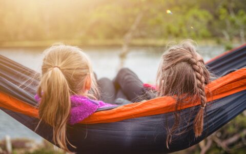 kaksi nuorta tyttöoletettua istuvat riippumatossa ja katsovat järvimaisemaa. Molemmilla henkilöillä on pitkät hiukset