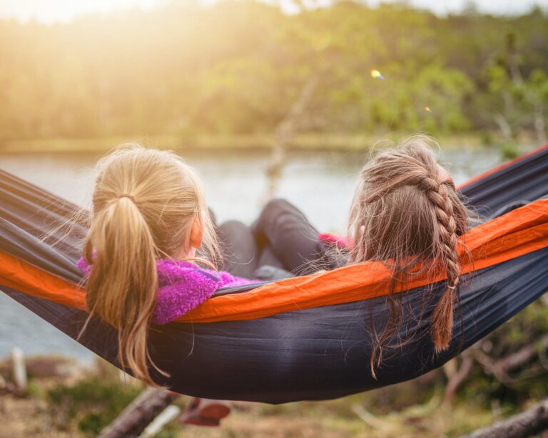 kaksi nuorta tyttöoletettua istuvat riippumatossa ja katsovat järvimaisemaa. Molemmilla henkilöillä on pitkät hiukset